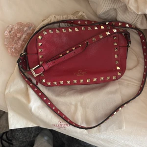 Röd axelväska Valentino Garavani - Snygg röd axelväska från Valentino Garavani i äkta skinn med coola nitar runt kanterna och på axelremmen. Väskan har en mjuk, rundad form och justerbar rem. Perfekt för dig som gillar statement-accessoarer och vill sticka ut med lyxig design. Pris kan diskuteras 