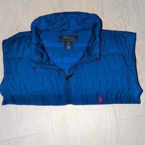 Blå Polo Ralph Lauren väst 14-16 stl - Snygg och klassisk dunväst från Polo Ralph Lauren, äkta såklart. Västen är i mycket fint skick - inga fläckar eller hål. Perfekt till höst, vår eller som lager under en jacka på vintern                             västenKungsblå med det ikoniska röda Polo-loggan på bröstet.  Har två fickor med dragkedja och en hög krage som håller värmen bra. -Kom med prisförslag!-