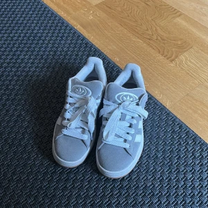 Adidas campus strl 36 - Säljer ett par grå Adidas sneakers i mocka med vita detaljer och klassiska tre ränder på sidan. Skorna har vit sula, rund tå och snörning. Adidas logga syns tydligt på plösen och insidan. Perfekta för dig som gillar stilrena och bekväma sneakers. Kartong medföljer. 