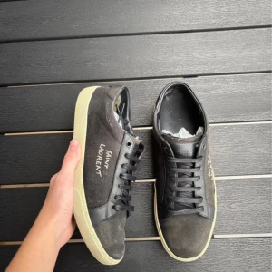 Svarta Saint Laurent sneakers - Svarta sneakers från Saint Laurent med vit textlogga på sidan. Skorna har svart snörning, rund tå och platt, beige sula. Ovandelen är i en mix av skinn och textil för en cool kontrast. Medföljer originalkartong och dustbag.