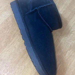 Svarta UGG boots i mocka - Mysiga svarta UGG boots i mocka med rund tå och platt sula. Insidan är fodrad med mjukt material för extra värme. Klassisk UGG-logga baktill och grov yttersula som ger bra grepp. Perfekta för kyliga dagar.