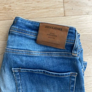 Blå jeans från Jack & Jones - Snygga blå jeans från Jack & Jones med klassisk femficksdesign och brunt läderpatch bak i midjan. Jeansen har en rak passform och är tillverkade i slitstarkt denimtyg. Perfekt för dig som gillar en enkel och tidlös stil.