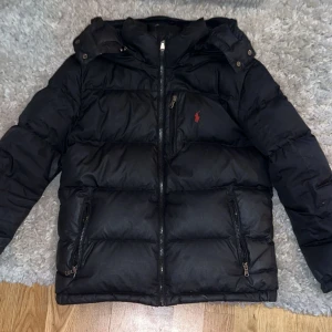 Svart pufferjacka från Polo Ralph Lauren - Säljer en svart pufferjacka från Polo Ralph Lauren med ikonisk röd logga på bröstet. Jackan har huva, två dragkedjefickor och kraftig vaddering som håller dig varm. Perfekt för kalla dagar och har en clean, stilren look.