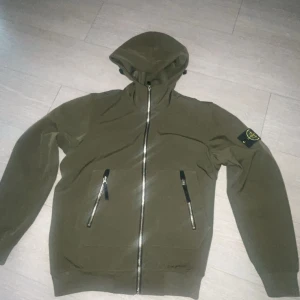 Olivgrön vindjacka från Stone Island - Säljer en olivgrön vindjacka från Stone Island då den inte kommer till användning längre. Jackan har en  klassisk patch på ärmen. Perfekt för dig som gillar streetwear och vill ha en clean look. 