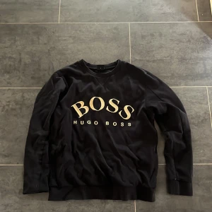 Svart sweatshirt från Hugo Boss S - Svart sweatshirt från Hugo Boss med stor BOSS-logga i guld på bröstet. Tröjan har rund hals, ribbade muddar och lång ärm. Materialet är mjukt och känns som bomull, perfekt för chill dagar. Snygg och enkel design som funkar till jeans eller joggers.