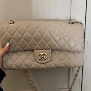 Beige axelväska - Superfin beige axelväska  i quiltat skinn med klassisk CC-logga i guld framtill. Väskan har kedjerem med skinn, innerfoder i vinrött och dragkedjefack under locket. Perfekt för dig som vill ha en ikonisk och lyxig väska med snygga detaljer.