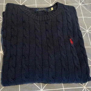 Mörkblå stickad tröja från Polo Ralph Lauren - Säljer en mörkblå stickad tröja från Polo Ralph Lauren med klassiskt kabelstickat mönster och röd broderad logga på bröstet. Tröjan har rund halsringning och långa ärmar, perfekt för kyligare dagar. Skick 8/10 med en defekt på qr koden av tvättning. Storlek S men passar M.
