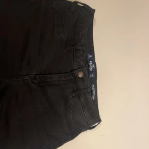 Svarta skinny jeans från Hollister - Svarta skinny jeans från Hollister med slitningar på benen för en edgy look. Klassisk ficksmodell med dragkedja och knapp framtill. Jeansen har hög midja och är tillverkade i stretchig bomullsblandning för skön passform.