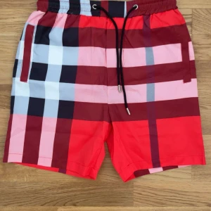 Röd-rutiga shorts från Burberry - Snygga shorts från Burberry med klassiskt rutigt mönster i rött, vinrött, svart, vitt och rosa. De har elastisk midja med svart snörning och sidofickor. Perfekta för sommaren och ger en riktigt fräsch vibe till din outfit.