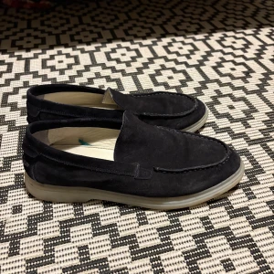 Svarta mockaloafers från Gant - Snygga svarta loafers från Gant i mjuk mocka med diskret logga på hälen. Skorna har en klassisk rund tå och platt sula i ljusgrått. Perfekta för dig som gillar stilrena och bekväma skor med en clean look.