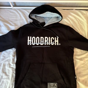 Svart hoodie från Hoodrich - Svart hoodie från Hoodrich med stor vit logga på både fram- och baksidan. Tröjan har huva med ljusgrå insida, snörning och en stor magficka. Perfekt för en streetwear-look och chill dagar.