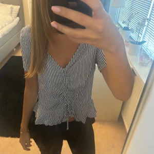 Randig blus med knyt och v-ringning - Säljer en blåvit randig blus med v-ringning och korta ärmar. Toppen har snygg snörning framtill som ger en rynkad effekt och knyt längst ner. Luftigt material och avslappnad passform, perfekt för varma dagar.