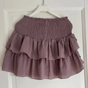 Ljuslila volangkjol med resår - Supersöt kort kjol i ljuslila med bred smock i midjan och dubbla volanger. Kjolen är luftig och har en feminin look som passar perfekt till sneakers eller sandaler. Tillverkad i ett tunt och mjukt material som känns skönt mot huden.