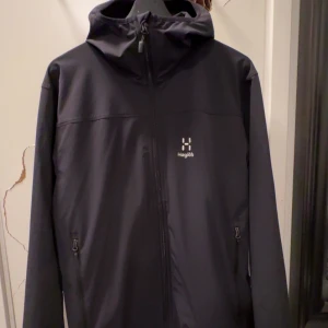 Svart vindjacka från Haglöfs med huva - Svart vindjacka från Haglöfs med minimalistisk design, hel dragkedja framtill och två sidofickor med dragkedja. Jackan har en fast huva och diskret Haglöfs-logga på bröstet. Perfekt för blåsiga dagar och outdoor-aktiviteter. Helt ny, använd 1 gång. 10/10 skick. Ny pris 2200kr