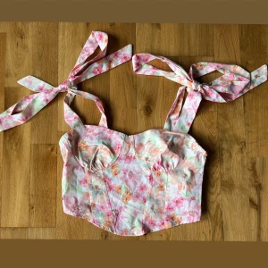 Blommig korsettopp med knytband - Supersöt korsettopp med blommigt mönster i rosa, orange och grönt. Axelbanden knyts i stora rosetter på axlarna för en extra gullig touch. Toppen har markerade kupor och figurnära passform, perfekt till sommarens outfits🌼🌸🌺