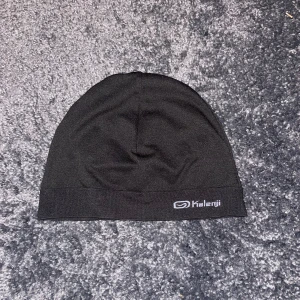 Svart träningsmössa från Kalenji - En stilren svart mössa från Kalenji, perfekt för löpning eller annan träning. Mössan är tillverkad i mjukt syntetmaterial som andas och har en diskret logga i vitt framtill. Enkel design med smal kant nedtill för bra passform.