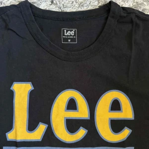 Svart Lee t-shirt med gul logga - Svart t-shirt från Lee i 100% bomull med stor gul och blå Lee-logga framtill. Klassisk rund hals och korta ärmar, perfekt för en chill och stilren look. Skön passform och mjukt material.
