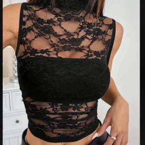 Svart spetsig croppad blus med hög hals - Säljer en svart croppad blus i spets med hög hals och ärmlös design. Toppen har transparenta partier med blommigt spetsmönster och en tight passform som framhäver figuren. Perfekt att styla med jeans eller kjol för en trendig look.