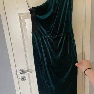 Mörkgrön sammetsklänning med spets - Snygg långklänning i mörkgrön sammet med en svart spetsdetalj vid ryggen. Klänningen har en oneshoulder-look och smal passform som ger en elegant siluett. 