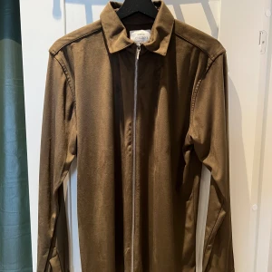 Brun mocka overshirt från Ciszere - Snygg brun overshirt från Ciszere i mjuk mocka-look. Jackan har klassisk krage, lång ärm och hel dragkedja framtill. Slim fit för en clean och stilren vibe, perfekt att slänga över en t-shirt eller hoodie.