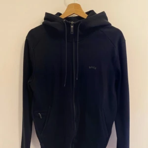 Svart hoodie från BOSS med dragkedja - Svart hoodie från BOSS med hel dragkedja framtill, snörning i huvan och diskret BOSS-logga på bröstet. Tröjan har långa ärmar och två fickor med dragkedja. Perfekt för en chill och stilren look. (Använt den 1 gång och köpte i Tyskland.) inga skador, hänger i garderoben har inte rört den 3 år. 