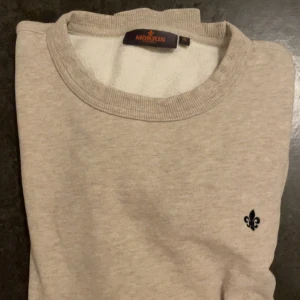 Beige sweatshirt från Morris, M - Snygg beige sweatshirt från Morris med rund hals och diskret svart lilje-logga på bröstet. Tröjan har ribbade muddar vid hals, ärmslut och nederkant. Perfekt för en clean och stilren look.