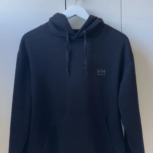 Svart Hov1 hoodie med tourtryck - Svart hoodie från Hov1 med liten krona broderad på bröstet och stort tourtryck på ryggen med alla turnédatum. Tröjan har huva med snörning och en stor magficka. Perfekt för dig som gillar svensk musik och vill ha en chill style