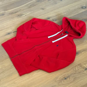 Röd hoodie från Polo Ralph Lauren - Snygg röd hoodie från Polo Ralph Lauren med vit snörning och dragkedja framtill. Tröjan har huva, långa ärmar och klassisk blå logga broderad på bröstet. Perfekt för chill dagar och enkel att matcha med jeans eller joggers.