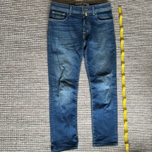 Blå jeans från Jacob Cohen - Snygga blå jeans från Jacob Cohen med klassisk femficksdesign och kontrastsömmar. Jeansen har en läderpatch med logga bak i midjan och subtila slitningar framtill. Modellen har rak passform och är tillverkad i mjukt denimtyg för en bekväm känsla.                                  Orginal pris: 5600kr                                                                   Längd: 105-110cm                                                                      Passar: 175-183cm
