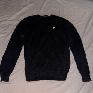 Svart stickad tröja från Lyle & Scott - Svart stickad tröja från Lyle & Scott med gul broderad logga på bröstet. Tröjan har ribbade muddar vid ärmslut och nederkant samt rund hals. Storlek S, pris kan diskuteras 