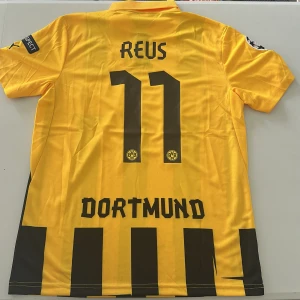 Borussia Dortmund Reus 11 matchtröja - Gul och svart Borussia Dortmund fotbollströja med Reus 11 på ryggen. Se bild 4, stjärnorna är inte centrerade, därav priset! Klassisk randig design, klubbmärke broderat på bröstet, Champions League-märke på ärmen och Evonik som sponsor. Tillverkad i lätt och ventilerande polyester. Replika, endast handtvätt gäller!