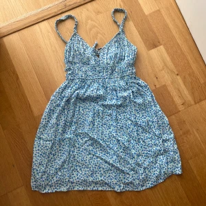 Blommig sommarklänning från Pull&Bear - Supersöt kort klänning från Pull&Bear med blått blommönster på vit botten. Smala axelband, med liten knytning framtill och smock i midjan. Perfekt för varma dagar och ger en fräsch och ungdomlig vibe.