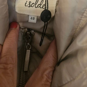 Beige quiltad jacka från Isolde - Snygg beige quiltad jacka från Isolde med dragkedja framtill och två fickor med dragkedja. Jackan har långärmade ärmar och en hög krage som ger en clean look. Perfekt för dig som gillar stilren och enkel design.