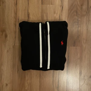 Svart hoodie från Polo Ralph Lauren - Svart hoodie från Polo Ralph Lauren med vit snörning, dragkedja och klassisk röd broderad logga på bröstet. Tröjan har huva, känguruficka och är tillverkad i mjuk bomull. Perfekt för chill dagar eller när du vill ha en clean streetstyle.