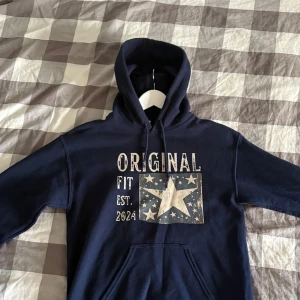 Mörkblå hoodie från Scndi  - Mörkblå hoodie från Scndi Original Fit med stor stjärna och texttryck framtill. Tröjan har huva med snörning och känguruficka. Tillverkad i en mjuk mix av bomull och polyester, perfekt för chill dagar. Snygg och enkel att matcha med jeans eller joggers.