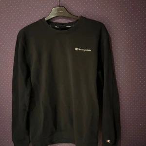 Svart sweatshirt från Champion - Svart sweatshirt från Champion i klassisk modell med rund hals och lång ärm. Diskret Champion-logga broderad på bröstet och vid ärmslutet. Tillverkad i mjuk bomullsmix som är skön att bära. Perfekt för en avslappnad och stilren look.