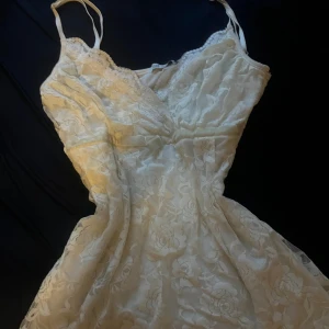 Vit spetstop/klänning  med blommönster - Superfin vit kortklänning i spets med blommigt mönster och smala axelband. Klänningen har v-ringad urringning och är figursydd upptill. Köpt secondhand. Använd några gånger 