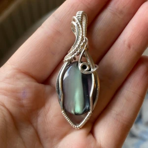 Silverberlock med norrskenssten - Säljer ett handgjord berlock med en oval, skimrande mörkgrön och svart sten infattad i detaljerad silvertråd. Hänget har en snygg, snirklig design som ger ett coolt och mystiskt intryck. Perfekt för dig som gillar unika smycken med lite edge.