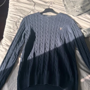 Mörkblå kabelstickad tröja från GANT - Snygg mörkblå kabelstickad tröja från GANT med rund hals och diskret logga på bröstet. Tröjan har långärmad passform och ribbade muddar vid hals, ärmslut och nederkant. Perfekt för dig som gillar klassisk stil med en modern touch.