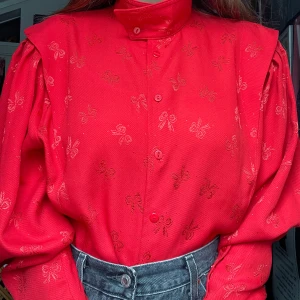 80-tals blus - Röd 80-tals blus med rosetter på mönstret. En krage som man kan vika ner eller ha uppe (se första och sista bilden). Det står att det är 38 i storlek men tycker den passar väldigt bra på mig med 36.  Jag kan mötas upp i Uddevalla centrum, annars fraktar jag gärna. Köparen står för frakten. (Står ej för postens slarv) katt finns i hemmet. 