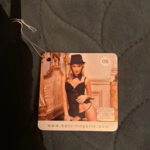 Svart babydoll-set från Baci Lingerie - Säljer ett svart babydoll-set från Baci Lingerie med tunna axelband, vadderade kupor och volangkant nertill. Setet inkluderar matchande stringtrosor. Materialet är mjukt och lätt, med små diskreta prickar på kuporna för extra detalj.