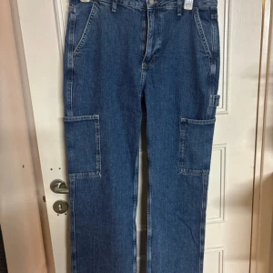 Blå cargobyxor i jeans, storlek M - Säljer ett par blå cargobyxor i jeansmaterial med raka ben och stora fickor på sidorna. Byxorna har klassisk femficksdesign, bälteshällor och är tillverkade i bomull. Perfekta för en avslappnad och trendig look.