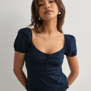 Mörkblå blus med puffärmar - Supersöt mörkblå blus med puffärmar och smockad rygg. Toppen har en djup, rundad urringning, små knappar framtill och volangkant nertill. Tillverkad i ett lätt, strukturerat tyg som känns skönt mot huden. 