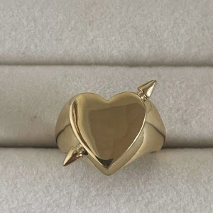 Guldig hjärtring med spjut - Cool ring i guldton med hjärtformad front och två spetsiga detaljer på sidorna. Ringen är öppen baktill vilket gör den justerbar. Perfekt statement-piece för dig som gillar unika accessoarer. Rostfri 