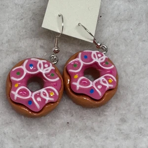 Donutörhängen med rosa glasyr - Supergulliga örhängen formade som donuts med rosa glasyr, vita slingor och färgglada strössel. Perfekt för dig som vill sticka ut och visa din lekfulla sida. Hängande modell med sterling silver krok. Nya