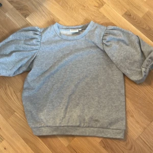 Grå tröja med puffärm från Kappahl - Grå tröja från Kappahl i mjuk bomullsmix med glittriga inslag. Tröjan har trendiga puffärmar och rund halsringning. Perfekt för dig som vill ha en enkel men ändå snygg look med en twist.