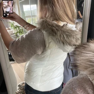 Vit dunväst med fuskpäls och stickad detalj - Säljer en snygg vit dunväst med dragkedja, stickad kabelmönstrad detalj upptill och huva med fluffig fuskpäls. Västen är quiltad och har fickor på sidorna. Perfekt för lager-på-lager och ger en fräsch look till din outfit.