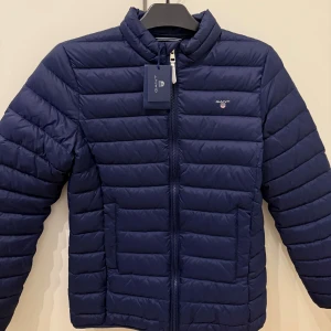 Marinblå pufferjacka från GANT - Snygg marinblå pufferjacka från GANT. Jackan har dragkedja framtill och två sidofickor. Perfekt för kyliga dagar. Storlek 176/ 16 år. Aldrig använd, lappar sitter kvar!