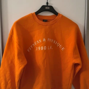 Orange sweatshirt från Gina Tricot XS - Cool orange sweatshirt från Gina Tricot i storlek XS. Tröjan har tryck med texten 'Fitness & Physique 1980 L.A.' framtill och är i mjuk bomull med ribbade muddar vid hals, ärmslut och nederkant. Perfekt för en sportig och avslappnad stil.