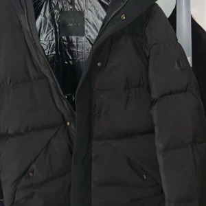 Svart Moncler dunjacka med huva - Säljer en svart dunjacka från Moncler med klassisk logga på ärmen. Jackan har quiltad design, dragkedja och tryckknappar framtill samt två fickor. Perfekt för kalla dagar och riktigt snygg streetstil.